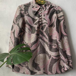Puede incluir: Una camisa de manga larga con botones y un estampado inspirado en el cachemir en tonos gris oscuro y rosa claro. La prenda cuenta con un cuello puntiagudo, una hilera vertical de botones forrados de tela y puños en las mangas.