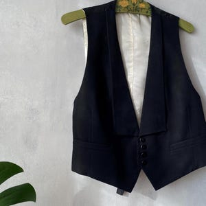 May include: A black vest featuring a deep V-neckline with wide lapels, a white interior lining, and a front closure secured by four small black buttons. The garment is sleeveless and includes two horizontal welt pockets on the lower front panels.