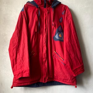 May include: A bright red hooded jacket featuring dark blue accents on the hood, shoulders, and hem. The front displays embroidered raindrop motifs in blue and orange, with two zipped pockets and a central zip closure with a protective flap.