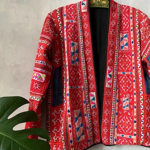 Könnte beinhalten: Eine langärmelige Jacke mit einer leuchtend roten Basis, verziert mit komplexen, mehrfarbigen geometrischen Stickereien und Patchwork-Einsätzen in Blau-, Rosa- und Weißtönen. Das vorne offene Kleidungsstück hat ein schwarzes Innenfutter.