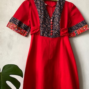 Pode incluir: Um vestido vermelho brilhante com mangas curtas e punhos estampados com desenhos florais em azul-marinho, vermelho e verde. O corpete inclui um painel floral azul-marinho com decote em V e uma fila vertical de cinco pequenos botões vermelhos.