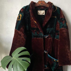 May include: A long-sleeved faux fur coat in deep brown, featuring a dark teal and yellow abstract pattern across the upper section. The coat has a shawl collar, toggle closures with teal trim, and a label inside the neck reading Veranica.