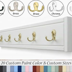May include: A white wooden coat rack with six brass hooks. The image shows a variety of hook finishes, including oiled bronze, brass, chrome, and white. The sign below indicates 20 custom paint colors and sizes are available.