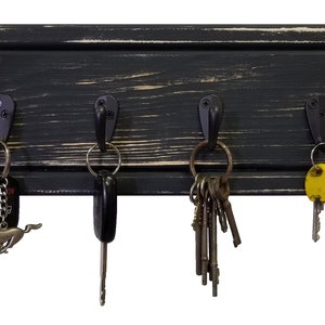 May include: A black wooden key holder with four metal hooks. The key holder has a distressed finish, with visible wood grain and a slightly worn appearance. Various keys hang from the hooks, including car keys and a key with a Mustang emblem.