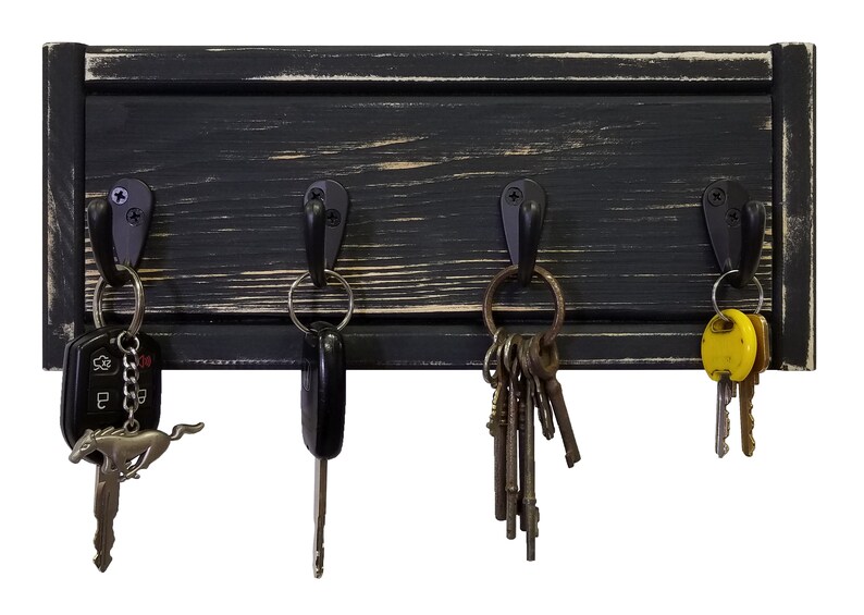 May include: A black wooden key holder with four metal hooks. The key holder has a distressed finish, with visible wood grain and a slightly worn appearance. Various keys hang from the hooks, including car keys and a key with a Mustang emblem.