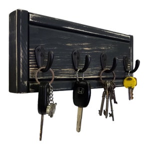 May include: A black wooden key holder with five metal hooks. The wood has a distressed finish, revealing some of the natural wood grain. Several sets of keys hang from the hooks, including car keys and vintage keys.