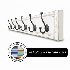 May include: A white wooden coat rack with seven black metal hooks. The rack has a distressed finish and is labeled "Handmade in Pennsylvania". The image also includes text that says "20 Colors & Custom Sizes".