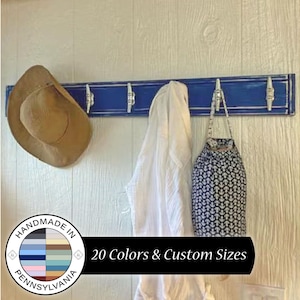 May include: A blue wooden coat rack with four hooks and a white distressed finish. The rack is mounted on a white wall. A brown cowboy hat, a white shirt, and a black and white patterned bag are hanging on the hooks. The text "Handmade in Pennsylvania" is on a circular sticker below the rack.