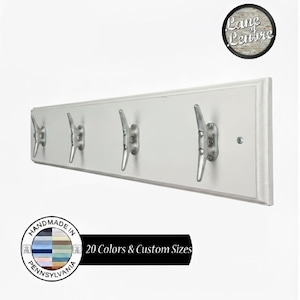 May include: A white wooden wall-mounted coat rack with five silver cleat-style hooks. The rack has a framed design and is handmade in Pennsylvania. The image includes a circular logo with the text "Lane Lenore" and a banner that reads "20 Colors & Custom Sizes."