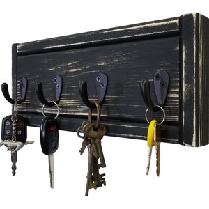 May include: A black wooden key holder with four metal hooks. The wood has a distressed finish with visible grain and white accents. Various keys and key fobs hang from the hooks, showcasing a practical and rustic design.
