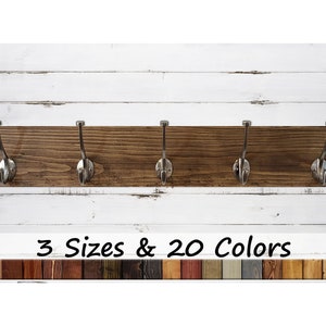 May include: A wooden coat rack with five hooks and a dark brown stain. The rack is mounted on a white wood wall. The text "3 Sizes & 20 Colors" is below the rack.