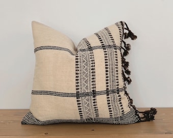 May include: A white pillow with black and brown geometric patterns and tassels. The pillow is on a wooden surface.