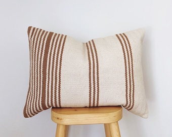 May include: A brown and white striped throw pillow sits on a wooden stool. The pillow is made of a soft, woven fabric and has a rectangular shape.