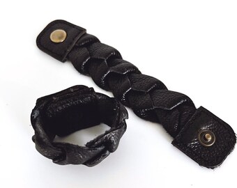 May include: A black leather bracelet with a braided design and a snap closure. The bracelet is made of two pieces, one that wraps around the wrist and another that snaps onto the first piece.