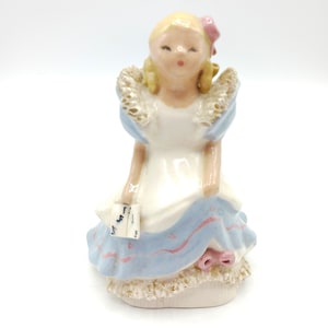 May include: A porcelain figurine of a girl wearing a blue and white dress with a pink bow. She has blonde hair and is holding a small book in her hand.