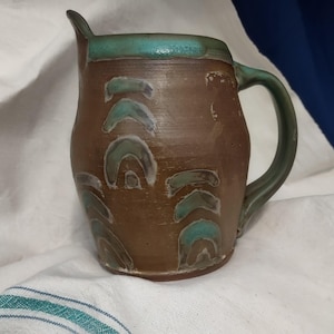May include: Handmade brown ceramic pitcher with a green handle and rim. The pitcher features a series of stylized, arch-shaped designs in white and teal. The pitcher has a spout for easy pouring and is a unique piece of pottery.