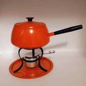 May include: An orange fondue pot with a black handle and lid. The pot sits on a black metal stand above a silver burner and an orange tray. The fondue pot is a classic design for entertaining.
