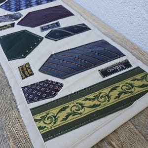 Könnte beinhalten: Ein rechteckiger Tischläufer aus cremefarbenem Stoff mit einer Collage aus verschiedenen Krawatten in Blau-, Grün-, Rot- und Violetttönen. Das Design enthält mehrere Krawattenetiketten und eine breite grüne Bordüre mit einem goldenen Blumenmuster.
