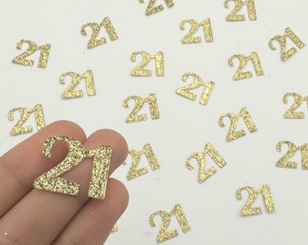 May include: Gold glitter number 21 embellishments scattered on a white surface. The numbers are small, with a textured, sparkly appearance. Ideal for crafting or party decorations, these numbers add a touch of glamour to any project.