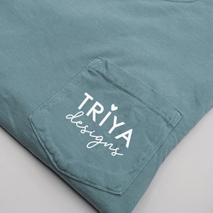 May include: A teal Comfort Colors pocket t-shirt. The pocket features the white text "TRIYA designs" with a heart symbol. The shirt is folded, showing the pocket and a portion of the crew neck collar.