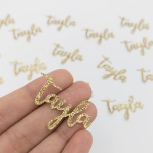 May include: A gold glitter name tag that says "Tayla" in a cursive font. The name tag is held in a hand, with several other blurred name tags in the background. The name tags are likely for crafts or personalized gifts.