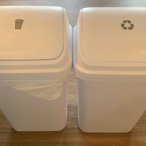 May include: Two white trash cans with lids. The left can has a trash can icon on the lid. The right can has a recycling symbol on the lid.