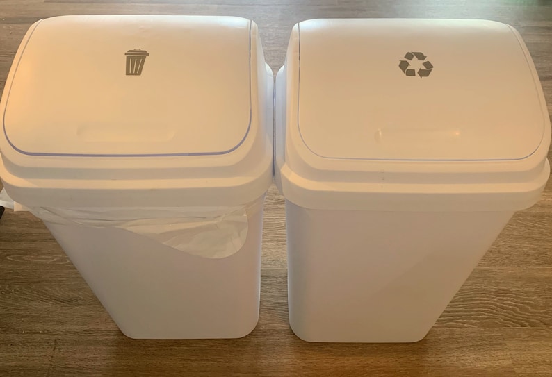 May include: Two white trash cans with lids. The left can has a trash can icon on the lid. The right can has a recycling symbol on the lid.