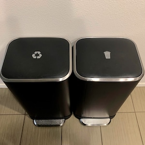 May include: Two black metal trash cans with silver lids and rims. The left can has a recycle symbol on the lid and the right can has a trash can symbol on the lid.