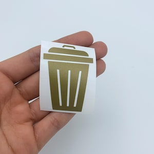 May include: Gold vinyl decal of a trash can with a lid and vertical lines on the side. The decal is on a white background.