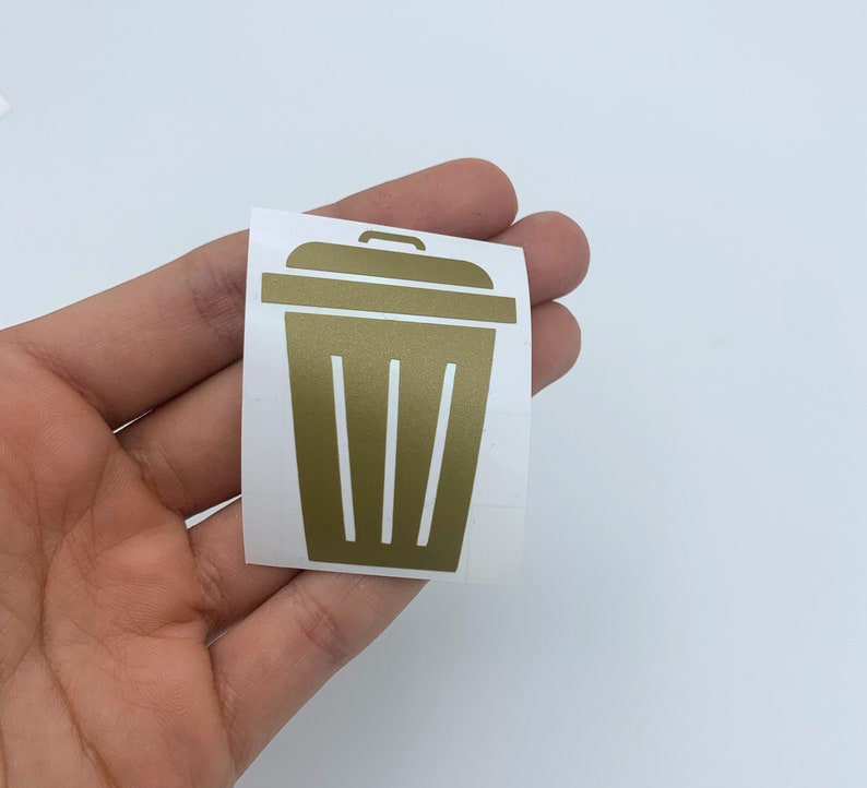 May include: Gold vinyl decal of a trash can with a lid and vertical lines on the side. The decal is on a white background.