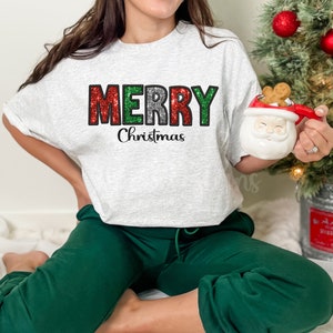 May include: A light gray sweatshirt with the words "MERRY Christmas" in red, green, and silver glitter lettering. The person is wearing green sweatpants. A Santa mug and Christmas tree are in the background.