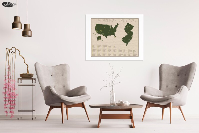 May include: A framed map of the United States with all the national parks and state parks highlighted in green. The map is on a beige background with a white frame. The map is hanging on a white wall in a living room setting with two armchairs and a coffee table.