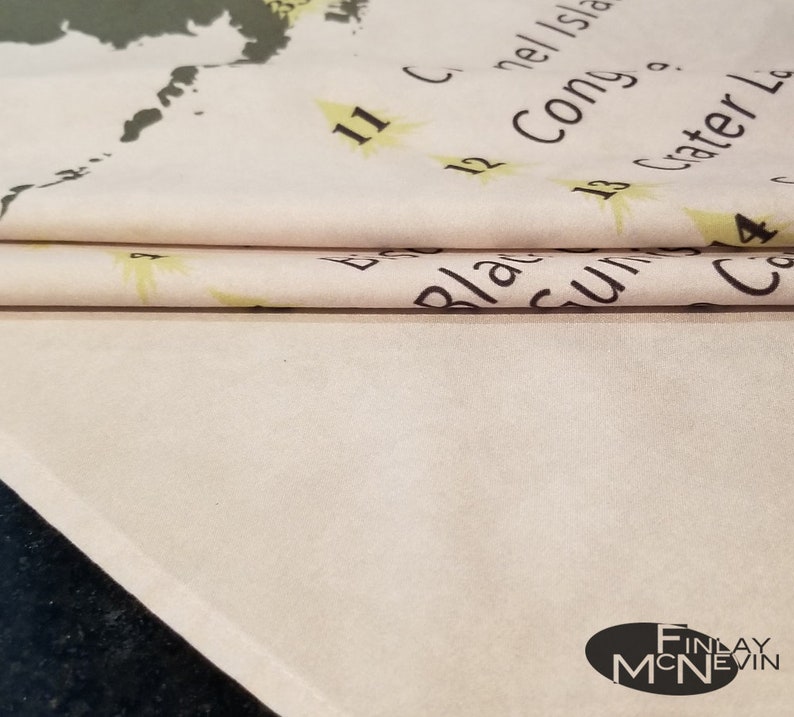 May include: A close-up of a folded white fabric with black text that reads "11 C. Connell Island", "12 Cong. o.", "13 Crater Lake", and "14 C", with a yellow star next to each number. The fabric has a light brown background and a white border.