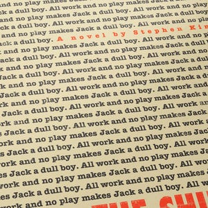 May include: A vintage book cover with a yellow background and black text. The title of the book is "The Shining" and the author is Stephen King. The text on the cover repeats the phrase "All work and no play makes Jack a dull boy" multiple times.