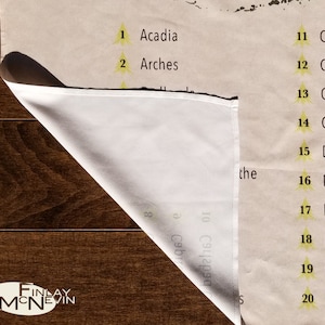 May include: A beige canvas print with a list of national parks in the United States. The list is numbered 1 through 20. The print is folded in half and the bottom corner is visible. The print is on a wooden surface.