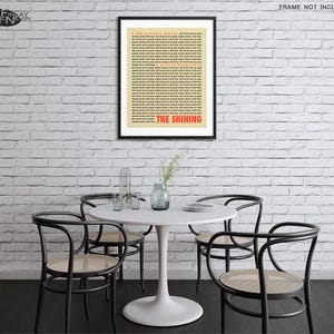 May include: A white brick wall with a black framed poster of the movie "The Shining" with a white background and red text. The poster is hanging on the wall above a white round table with four black chairs. There are plants on the table.