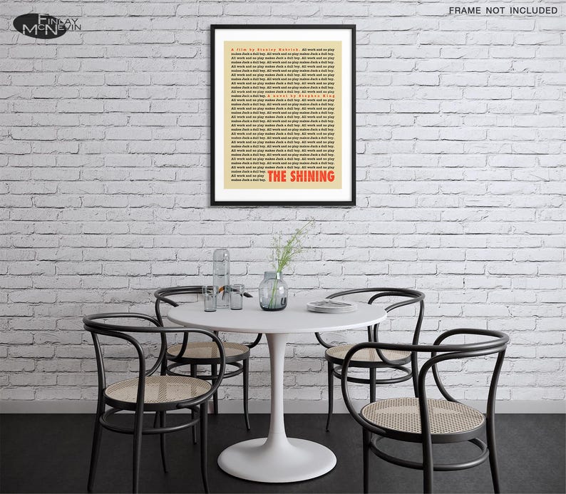 May include: A white brick wall with a black framed poster of the movie "The Shining" with a white background and red text. The poster is hanging on the wall above a white round table with four black chairs. There are plants on the table.