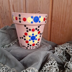 Pode incluir: Um vaso de flores cor de terracota com um design pintado à mão. O vaso apresenta um padrão de pontos coloridos em vermelho, amarelo, azul e preto. As palavras "San 1776" estão escritas no vaso. O vaso está sobre um tecido cinzento.