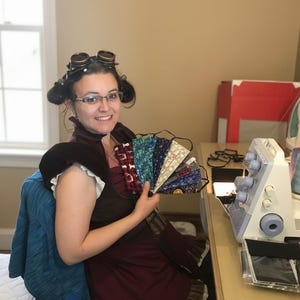 May include: A woman holding a fan of various fabric face masks. The masks display different patterns and colors, including floral, geometric, and abstract designs. A sewing machine is visible on a desk in the background.