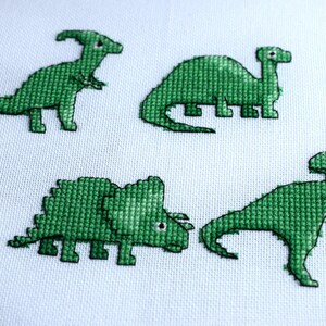 May include: Four green dinosaur cross-stitch designs on a white fabric background. The dinosaurs include a Parasaurolophus, a Brontosaurus, a Triceratops, and a Tyrannosaurus Rex. The cross-stitch is detailed and the dinosaurs are in various poses.