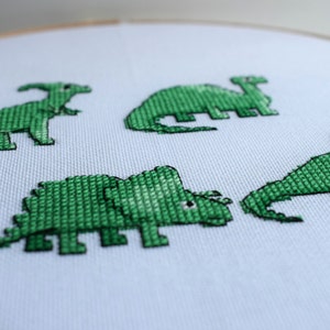 May include: A close-up of a white fabric stretched in a wooden embroidery hoop. Four green cross-stitched dinosaurs are displayed on the fabric. The dinosaurs include a Parasaurolophus, a Stegosaurus, a Triceratops, and a Brontosaurus.