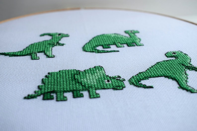 May include: A close-up of a white fabric stretched in a wooden embroidery hoop. Four green cross-stitched dinosaurs are displayed on the fabric. The dinosaurs include a Parasaurolophus, a Stegosaurus, a Triceratops, and a Brontosaurus.