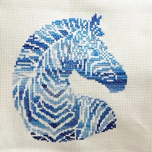 May include: A cross-stitch design of a horse head in shades of blue on a white background. The horse head is filled with geometric patterns and varying shades of blue thread, creating a textured appearance. The design is centered on the fabric.