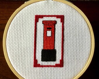 May include: A cross-stitch embroidery of a red British post box, with a black base and a white slot, set against a white background. The design is framed in a wooden embroidery hoop.