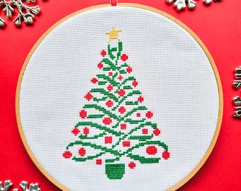 May include: A cross-stitch Christmas tree design in a wooden embroidery hoop. The tree is green with red ornaments and a yellow star on top, set against a white background. Silver snowflake ornaments are scattered around the hoop on a red surface.