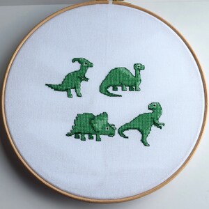 May include: A cross-stitch embroidery hoop featuring four green dinosaurs on white fabric. The dinosaurs include a Parasaurolophus, a Brontosaurus, a Triceratops, and a Tyrannosaurus Rex. The wooden hoop holds the fabric taut.