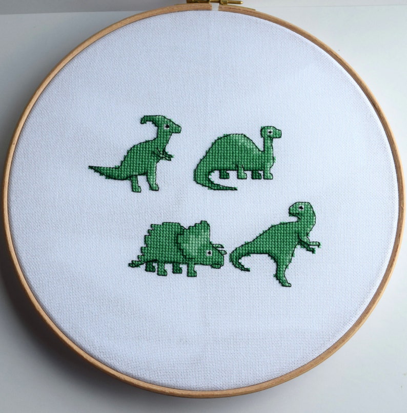 May include: A cross-stitch embroidery hoop featuring four green dinosaurs on white fabric. The dinosaurs include a Parasaurolophus, a Brontosaurus, a Triceratops, and a Tyrannosaurus Rex. The wooden hoop holds the fabric taut.