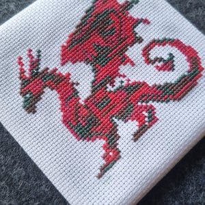 May include: A cross-stitch dragon design in red, green, and brown on white fabric. The dragon is displayed on a small, square piece of fabric, held by two wooden dowels. The background is a dark gray, textured surface.