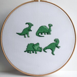 May include: A cross-stitch embroidery hoop featuring four green dinosaur designs on white fabric. The dinosaurs are in various poses, and the hoop is made of wood. The design is simple and charming.