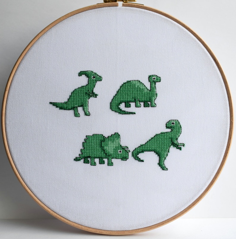 May include: A cross-stitch embroidery hoop featuring four green dinosaur designs on white fabric. The dinosaurs are in various poses, and the hoop is made of wood. The design is simple and charming.